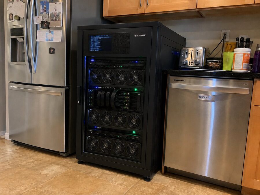 A black server rack full of fans and GPUs standing in a home kitchen between the fridge and the dishwasher